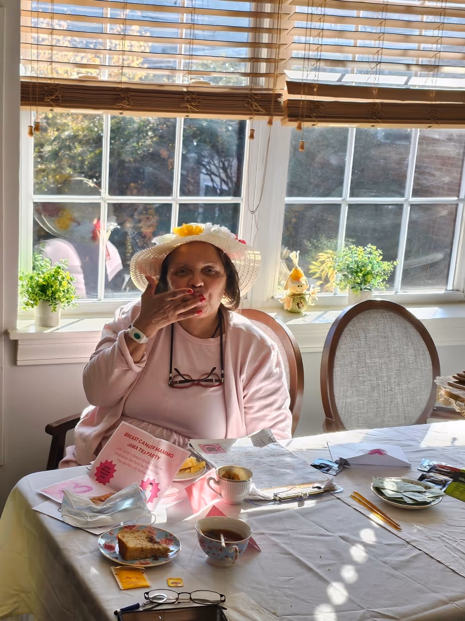 An older woman wearing a decorative sun hat sits at a table by a sunny window, blowing a kiss with tea cups, snacks, and papers on the table.