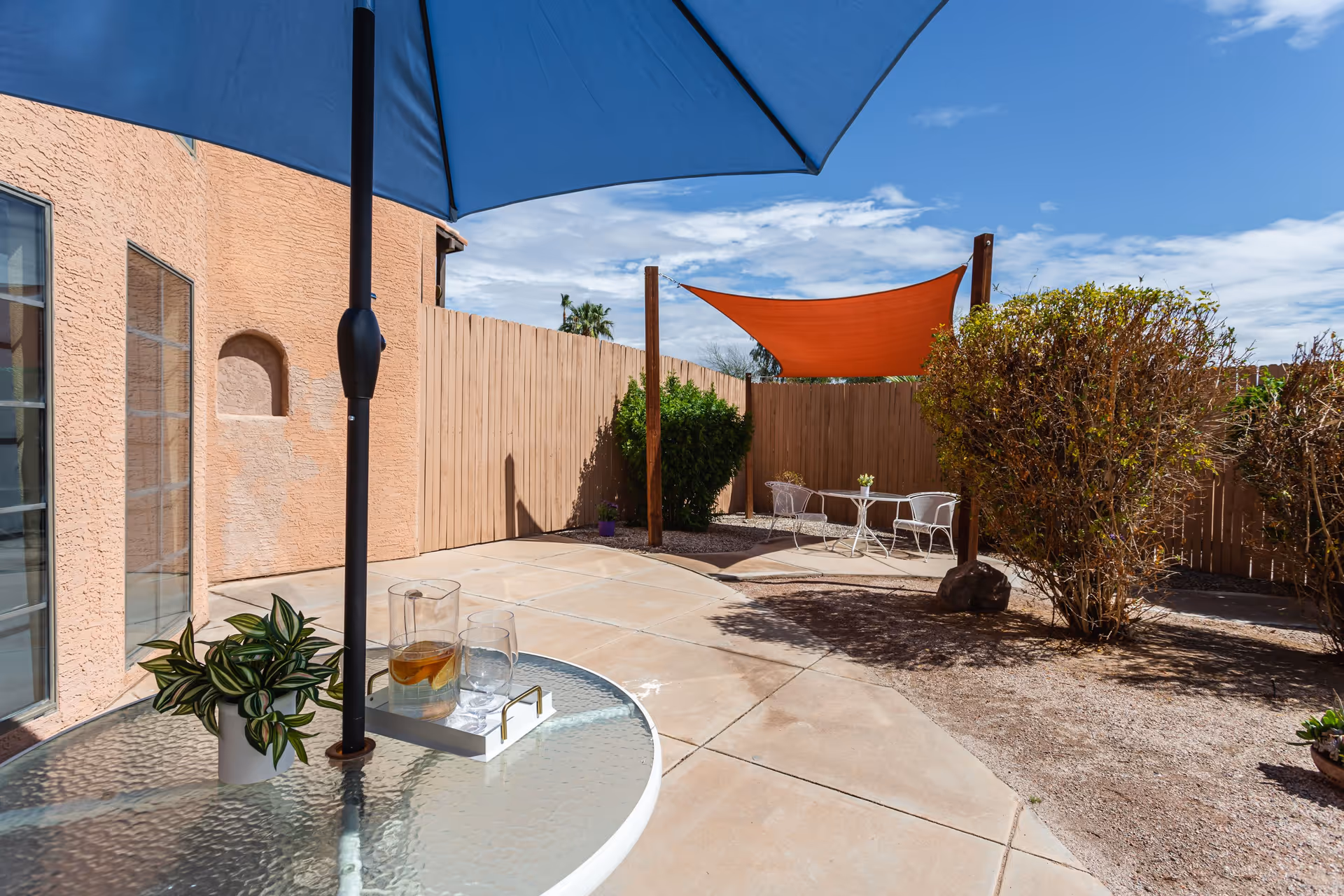 Outdoor patio area with a glass table under a blue umbrella, holding a pitcher of iced tea, a glass, and a small potted plant. In the background, there is a wooden fence, some bushes, and a small seating area with two white chairs and a table under an orange shade sail.