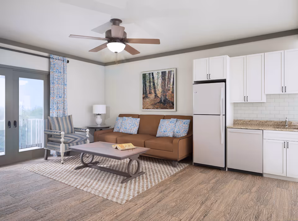 A bright living room and kitchen area with a brown sofa adorned with patterned blue and white pillows, a wooden coffee table on a textured rug, a striped armchair next to a side table with a lamp, white kitchen cabinets, a refrigerator, dishwasher, and a ceiling fan with light. There is a large window with blue patterned curtains and a framed painting of a forest on the wall.