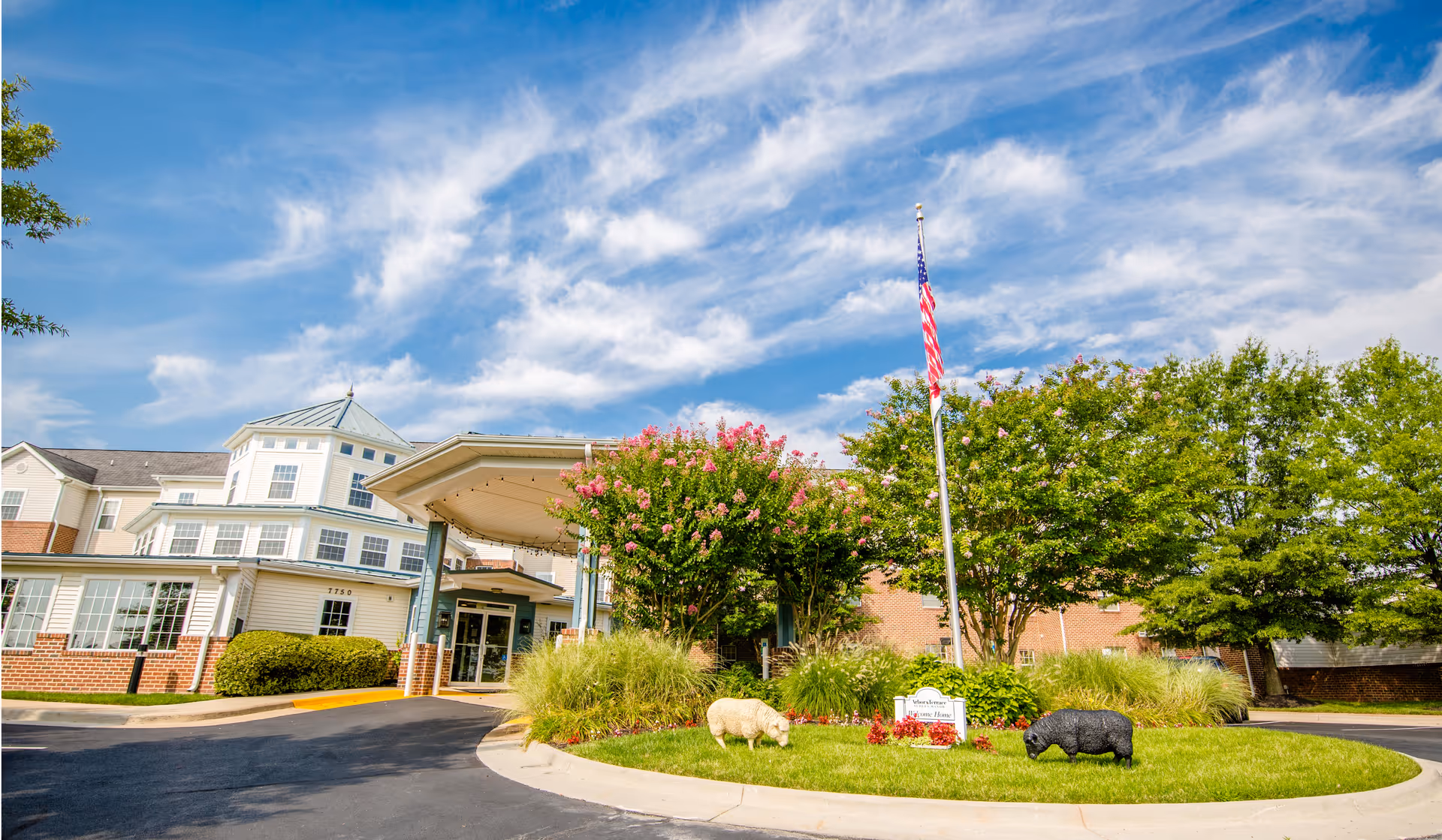 Exterior view of Arbor Terrace Sudley Manor senior living facility with a circular driveway, landscaped greenery, two decorative animal statues, an American flag on a flagpole, and a partly cloudy blue sky.