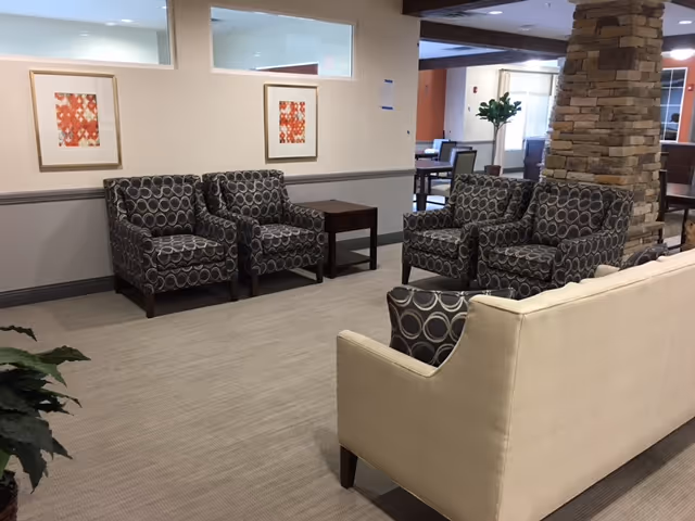 A seating area in a senior living facility with four patterned armchairs arranged around a small wooden table, a beige sofa with a patterned cushion, a stone pillar, and framed artwork on the wall. The space has carpeted flooring and neutral-colored walls.