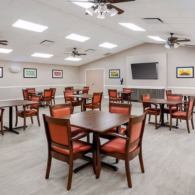 Spacious dining room with several wooden tables and red-upholstered chairs, ceiling fans, a wall-mounted TV, and framed artwork on the walls.