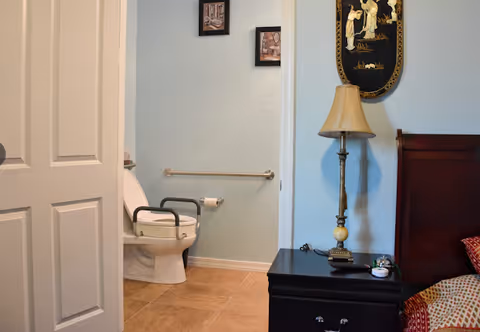 View of a bathroom with a toilet equipped with safety rails, partially visible through an open door from an adjacent bedroom. The bedroom has a dark wooden nightstand with a lamp and a bed with a patterned pillow. The bathroom wall has a grab bar and two small framed pictures.