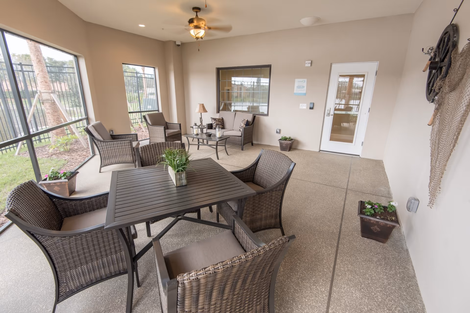 A covered outdoor patio area with wicker furniture including a square table with four chairs, a loveseat, two armchairs, and a coffee table. The patio has a ceiling fan with a light, potted plants, and large screened windows overlooking a fenced garden area with palm trees.