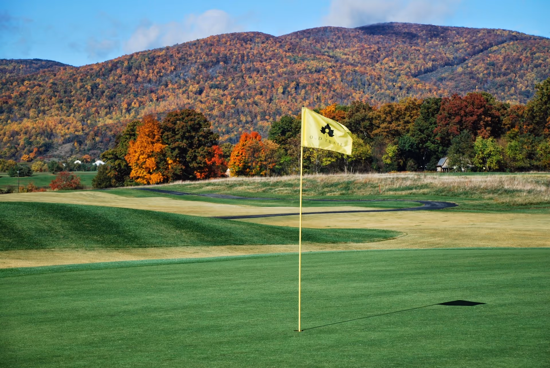 A golf course with a yellow flag marked 'Old Trail' on a putting green, surrounded by rolling hills and trees with autumn foliage under a partly cloudy sky.