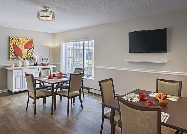 A dining area in a senior living facility with two wooden tables, each surrounded by four chairs. The tables are set with red cups, plates, and small decorative items. A large window lets in natural light, and a flat-screen TV is mounted on the beige wall above a small white shelf. A white sideboard with decorative items and a colorful painting is against one wall. The floor is wooden, and the room has a modern ceiling light fixture.