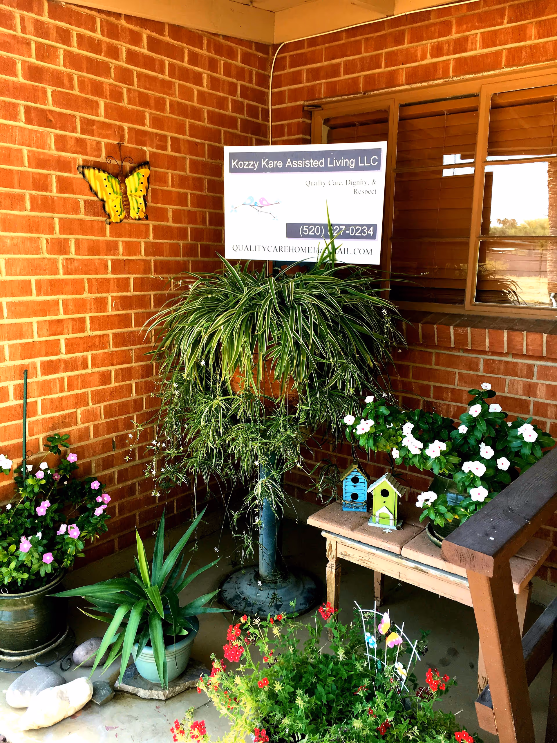 A cozy outdoor corner with various potted plants and flowers arranged around a small wooden table and a bench. A large leafy plant stands on a pedestal, and two small decorative birdhouses sit on the table. The background features a brick wall with a yellow butterfly decoration and a window. A sign on the wall reads 'Kozzy Kare Assisted Living LLC' with contact information.