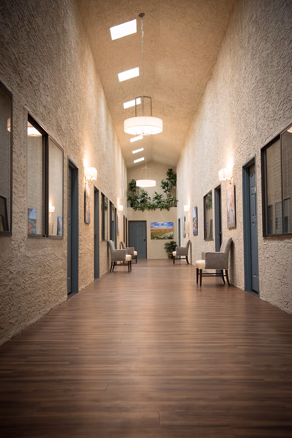 A long, narrow hallway with textured beige walls and a wooden floor. The ceiling is vaulted with several rectangular skylights and modern hanging light fixtures. Along the walls, there are multiple doors and windows, with framed artwork and wall sconces providing additional lighting. Several cushioned chairs are placed against the walls, and there are green plants at the far end of the hallway.