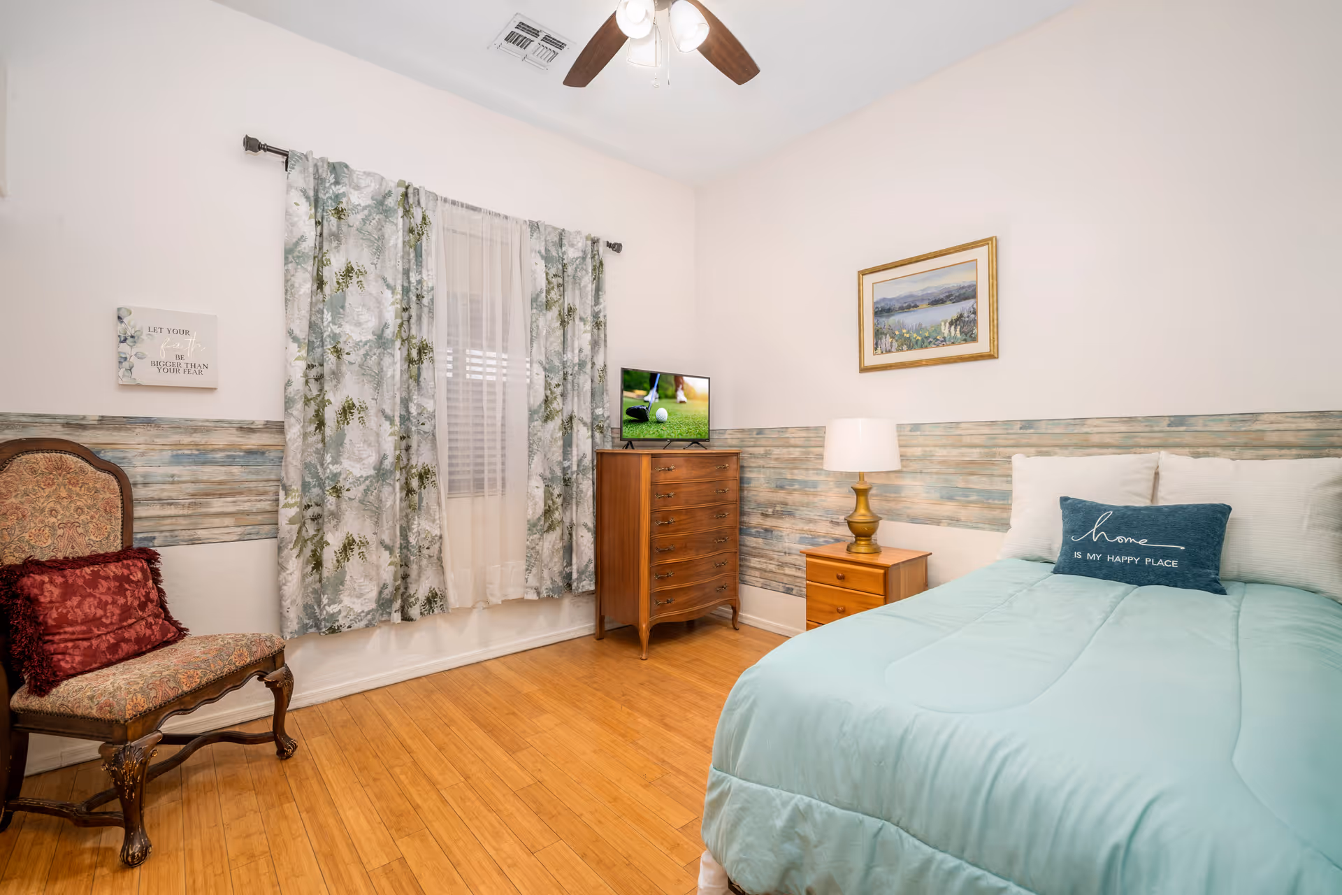 A cozy bedroom with a light blue bedspread on a bed, a decorative pillow that says 'home is my happy place', a wooden nightstand with a lamp, a wooden dresser with a small TV on top, a window with floral curtains, a framed landscape painting on the wall, and an upholstered chair with a red cushion.