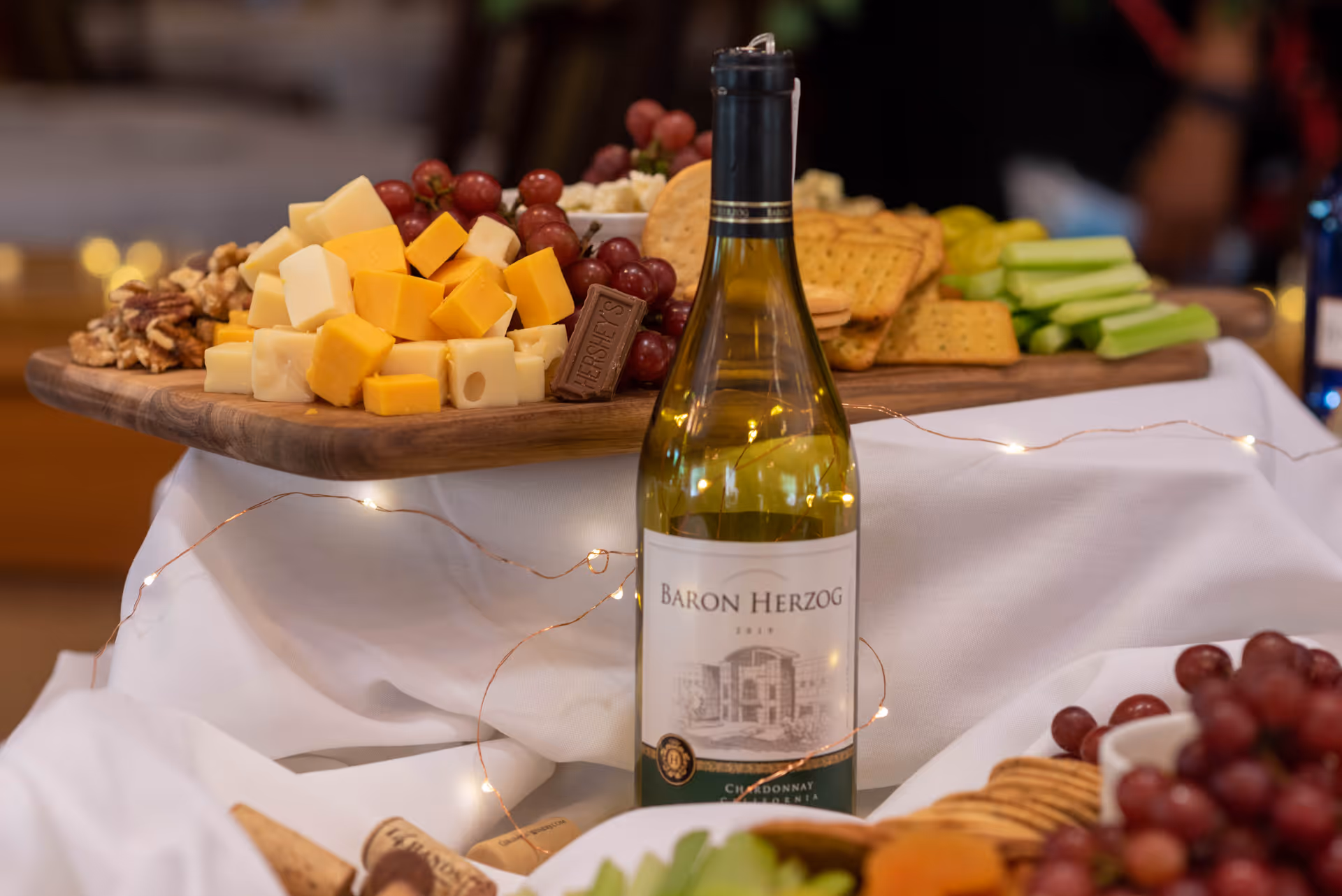 A close-up view of a charcuterie board featuring cubes of cheddar and Swiss cheese, grapes, crackers, celery sticks, nuts, and a piece of Hershey's chocolate. In front of the board is a bottle of Baron Herzog Chardonnay wine, surrounded by decorative string lights and a white cloth.