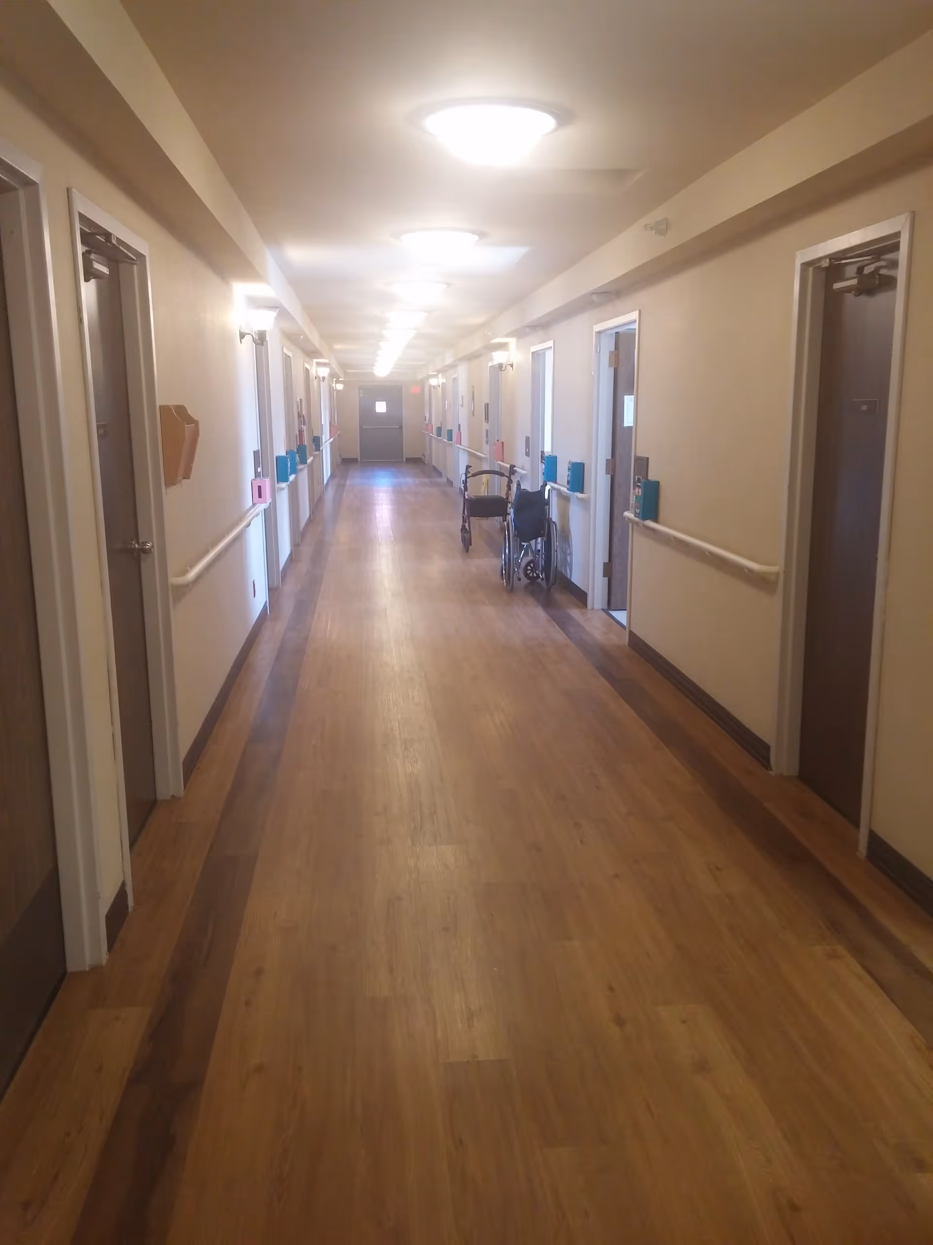 Long, well-lit interior hallway with wood-look flooring, doors and handrails on both sides and a wheelchair parked along the wall.