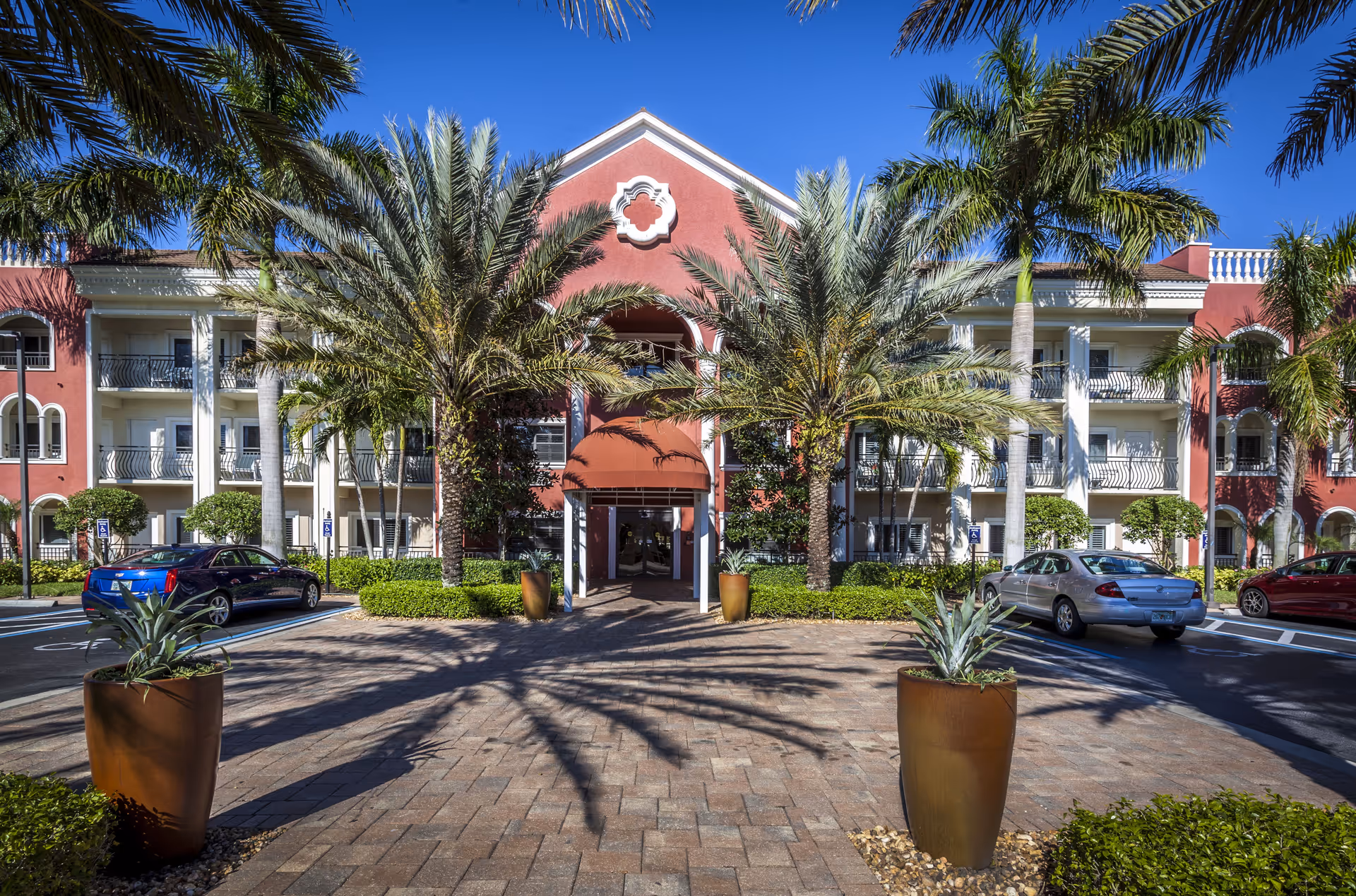 Front exterior view of Tuscany Villa of Naples, a multi-story building with white balconies and red walls, surrounded by palm trees and parked cars under a clear blue sky.
