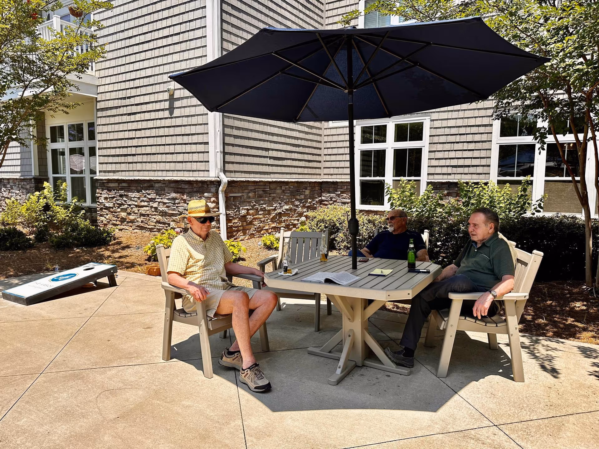 Three elderly men sitting around a patio table with a large black umbrella, outside a building with stone and shingle siding. There is a cornhole game set up on the concrete patio, and greenery including bushes and trees surrounding the area.