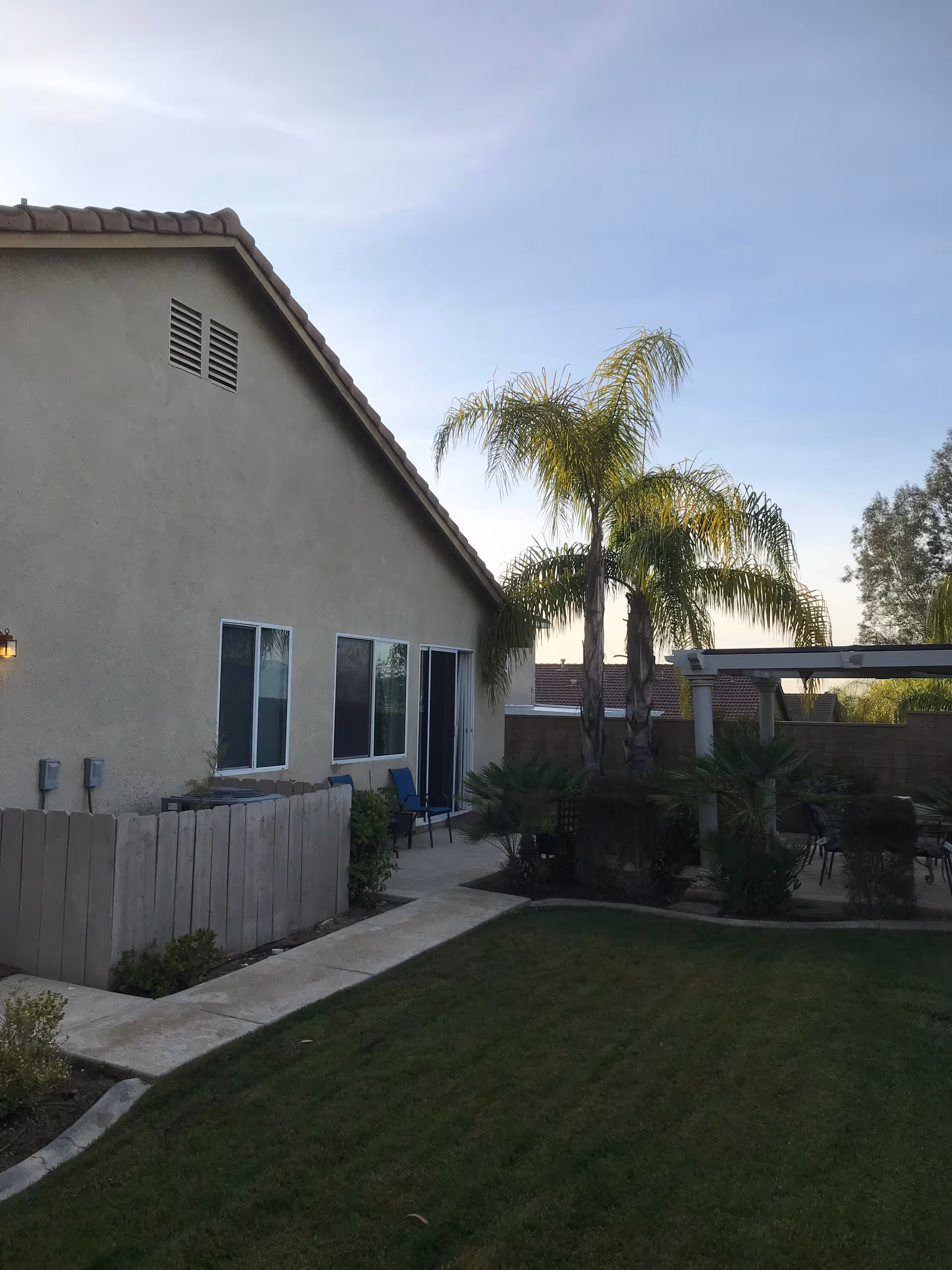 Backyard view of a single-story house with beige stucco walls and a tiled roof. There is a small concrete pathway along the side of the house, a wooden fence, green lawn, palm trees, and a covered patio area with outdoor furniture.