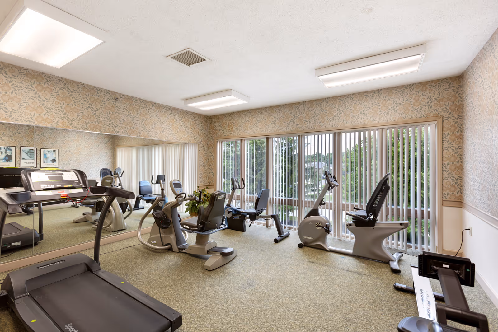 A fitness room with exercise equipment including a treadmill, stationary bikes, and a rowing machine. The room has large windows with vertical blinds allowing natural light to enter, floral wallpaper, and a large mirror on one wall reflecting the equipment.