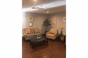 A cozy living room area with a gray sofa adorned with striped cushions, a beige armchair, a dark wooden coffee table, and a side table with a vase of yellow flowers. The walls are painted light gray and decorated with a large clock, framed pictures, and a tall green plant in the corner. The floor is dark wood.