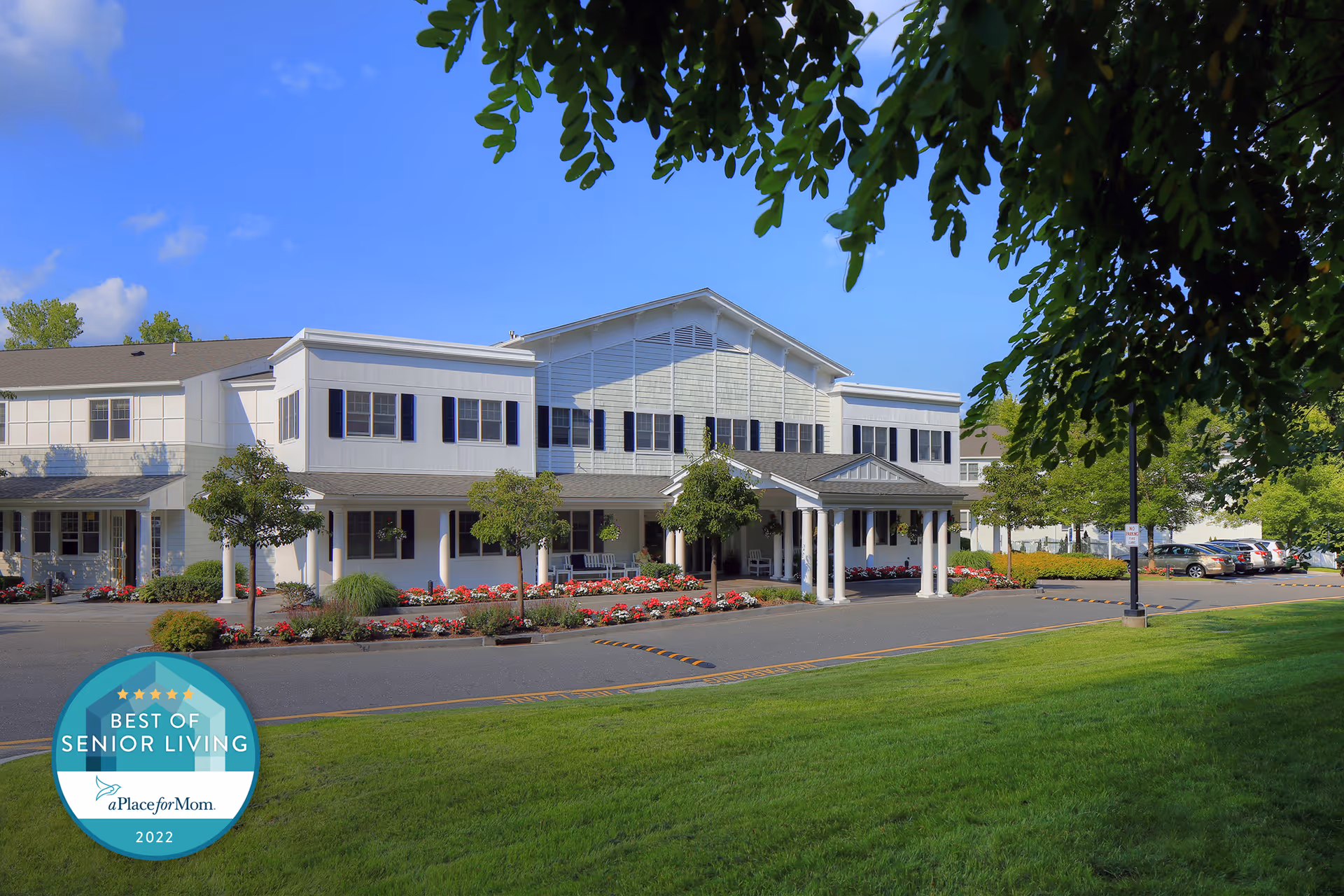 Exterior view of a senior living facility building with white siding, multiple windows, and a covered entrance. The building is surrounded by well-maintained landscaping including green grass, trees, and flower beds. Several cars are parked in the parking lot to the right. The sky is clear and blue.