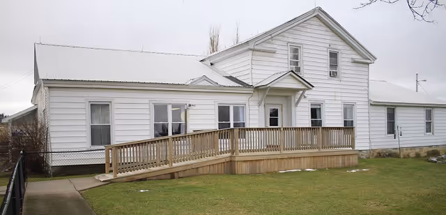 Exterior view of a white two-story building with a metal roof and a wooden wheelchair ramp leading to the entrance. The building is surrounded by a grassy lawn and a concrete walkway, with a chain-link fence on the left side.
