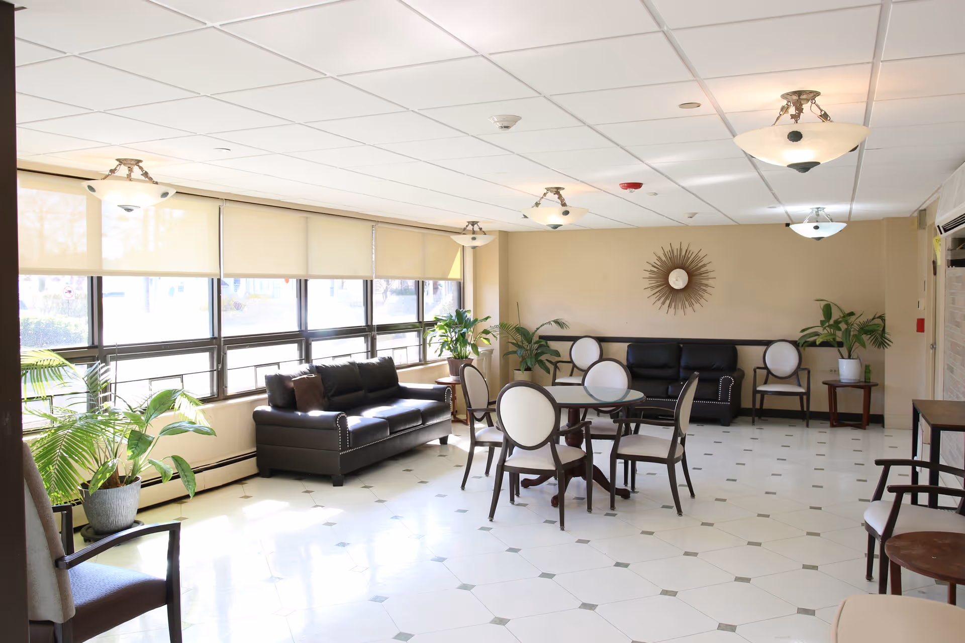 A bright and spacious common area with large windows covered by beige roller shades. The room features two black leather sofas, several round tables with white cushioned chairs, potted plants, and a decorative sunburst wall clock. The floor is tiled with a white and gray pattern, and ceiling lights provide additional illumination.