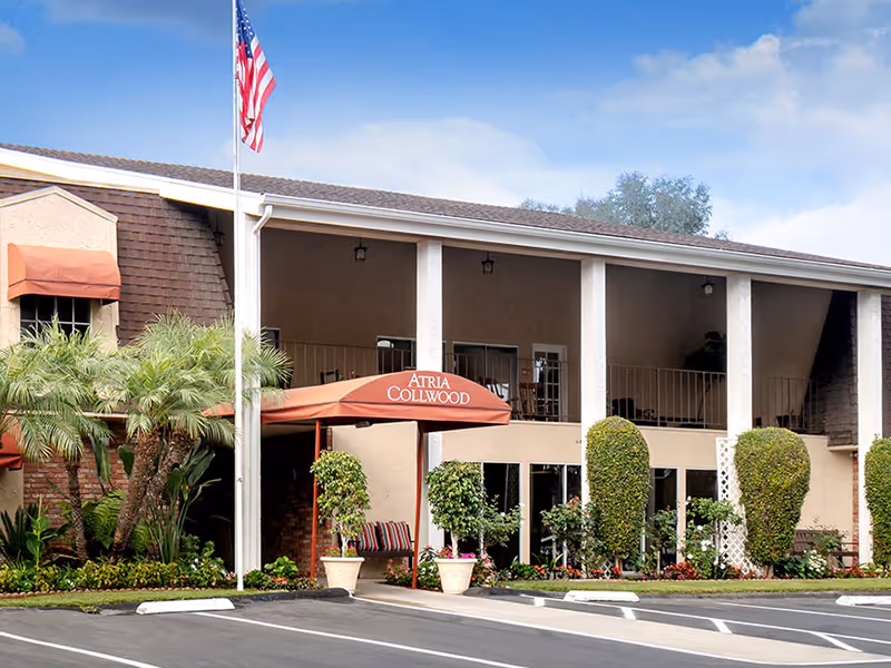 Exterior view of Atria Collwood senior living facility showing a two-story building with a covered entrance, potted plants, trimmed bushes, an American flag on a flagpole, and a clear blue sky.