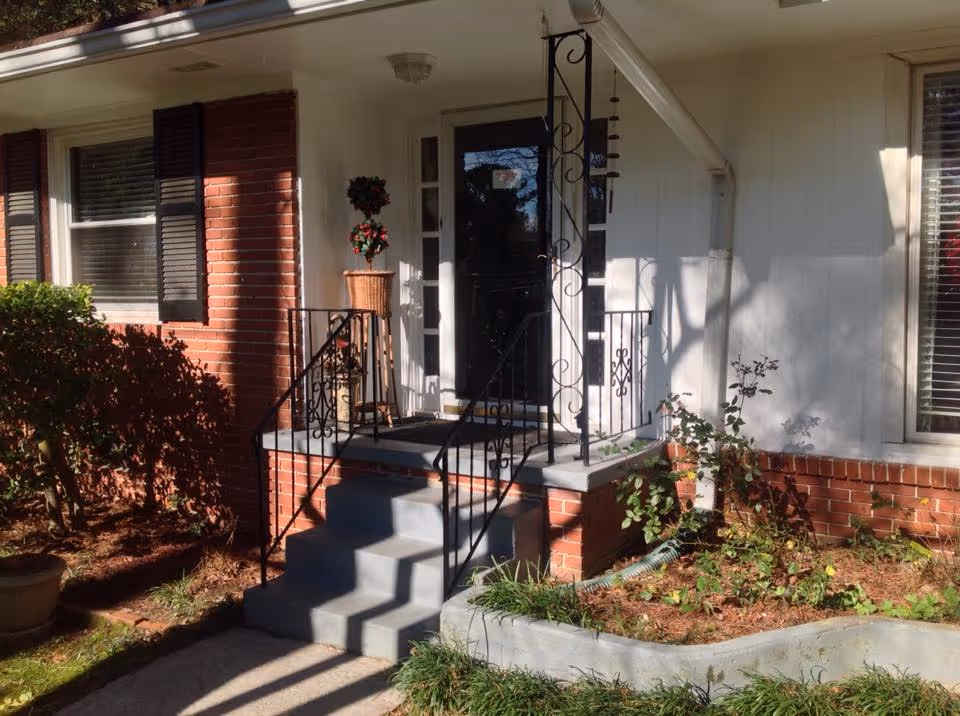 Front entrance of a brick house with concrete steps, decorative wrought-iron railings, a glass storm door, and potted plants.
