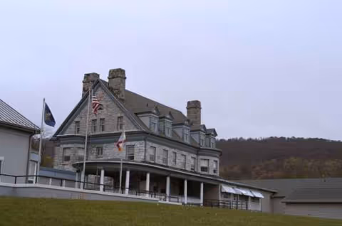 Stone mansion-style senior living facility exterior with flags, porch, and hillside backdrop.