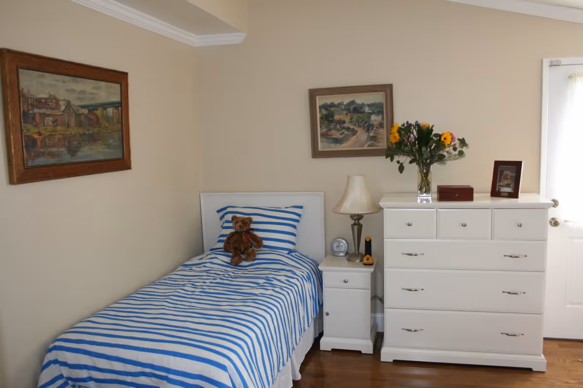 A small bedroom with a single bed covered in blue and white striped bedding, a teddy bear sitting on the bed, a white nightstand with a lamp and clock, a white dresser with flowers in a vase, a framed photo, and a small box on top. Two framed paintings hang on the beige walls.