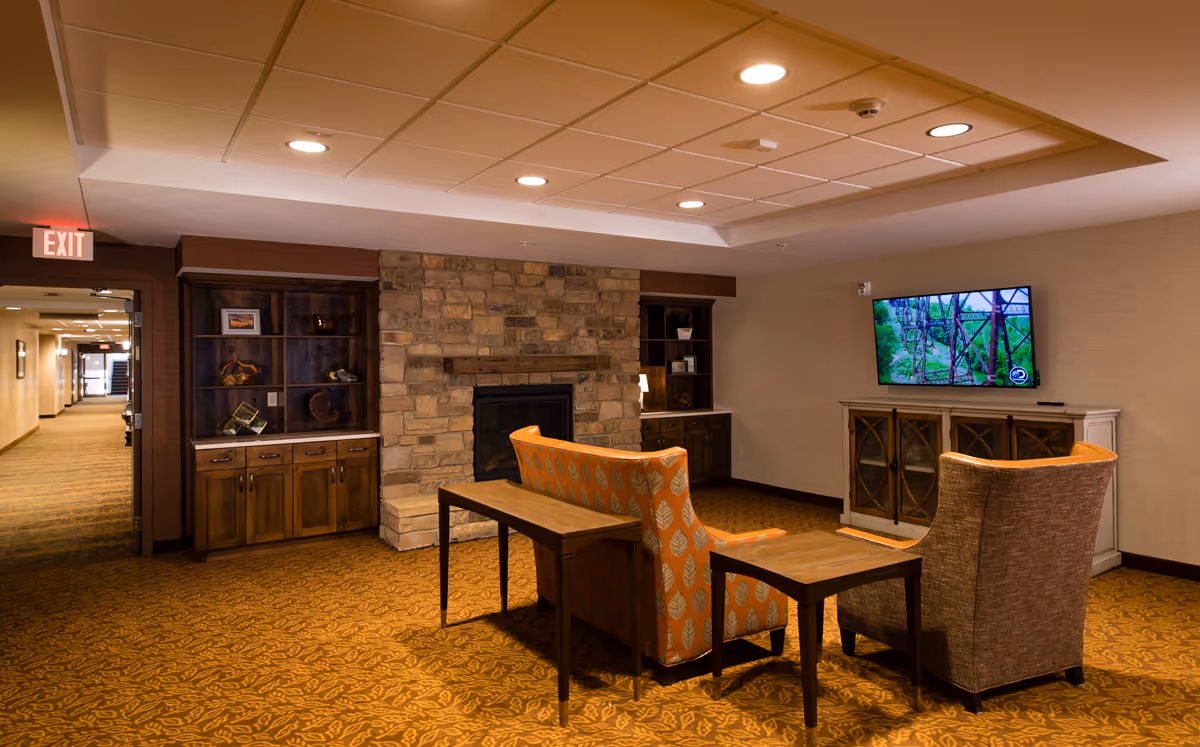 Cozy communal lounge with two patterned armchairs and a bench facing a wall-mounted TV, a stone fireplace, built-in cabinets, and patterned carpet.