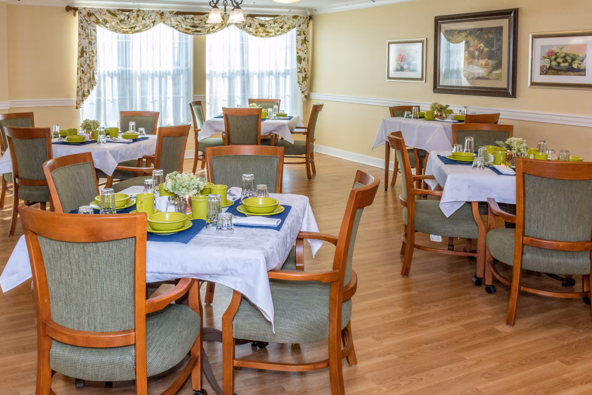 A bright dining room with several wooden tables and chairs arranged neatly. Each table is covered with a white tablecloth and set with green plates, bowls, cups, and clear glasses. The room has large windows with floral curtains allowing natural light to fill the space. The walls are painted light yellow and decorated with framed artwork.