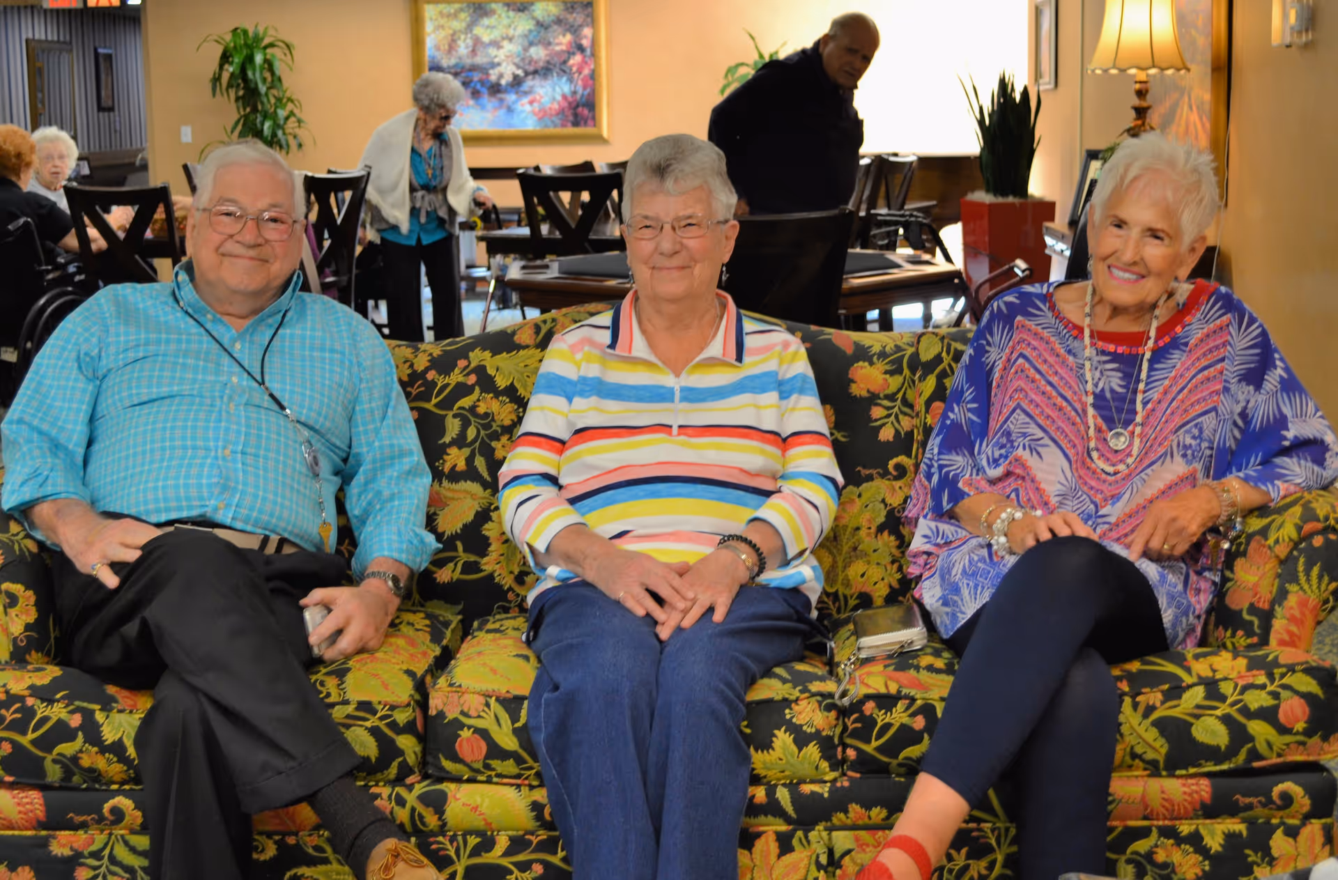 Three elderly people sitting on a floral patterned couch in a cozy living room or common area. The man on the left is wearing a blue checkered shirt and glasses, the woman in the middle is wearing a colorful striped shirt, and the woman on the right is wearing a purple patterned top with jewelry. In the background, other elderly people are visible, along with chairs, tables, plants, and a lamp.