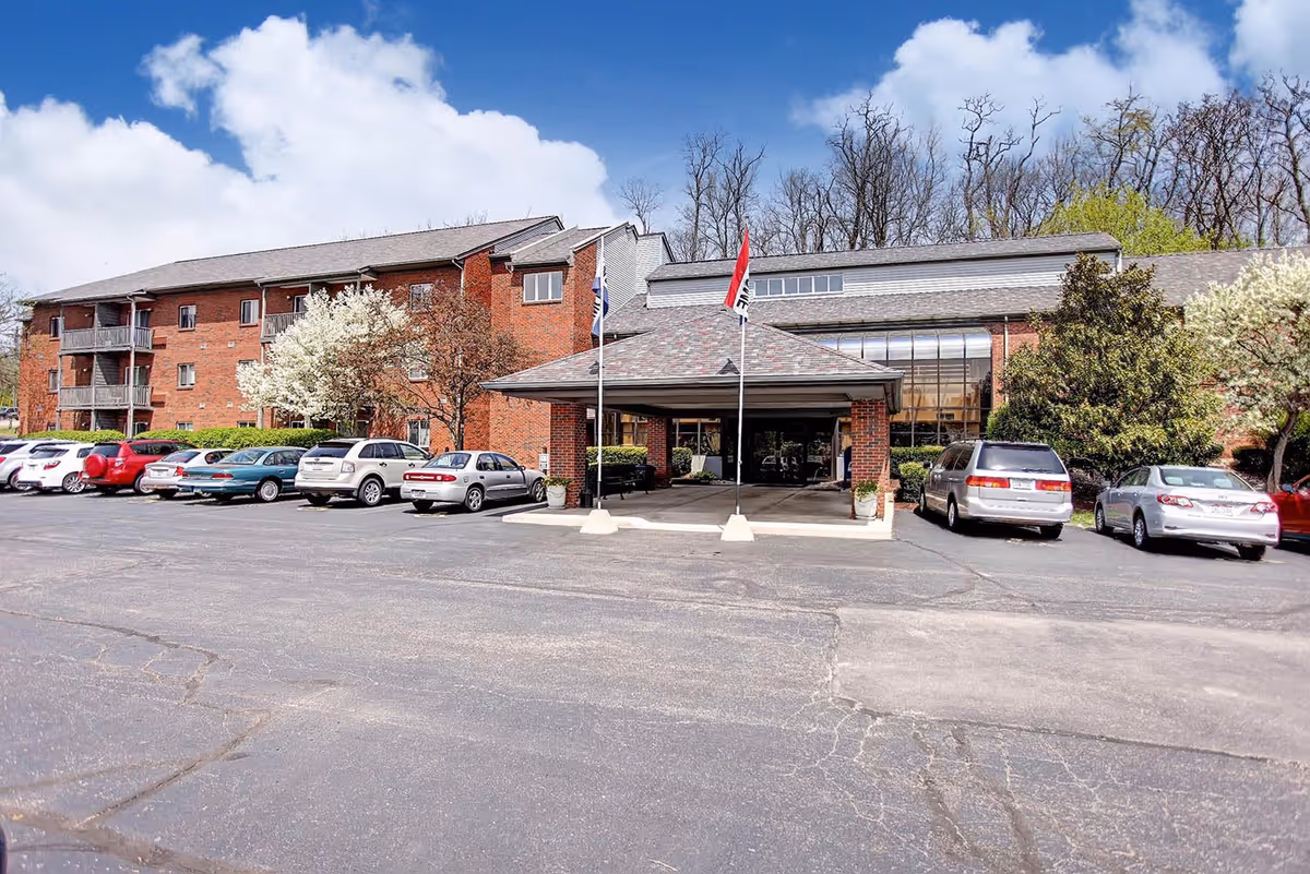 Front entrance of a brick senior living building with a covered drop-off, flagpoles, parked cars, and flowering trees.