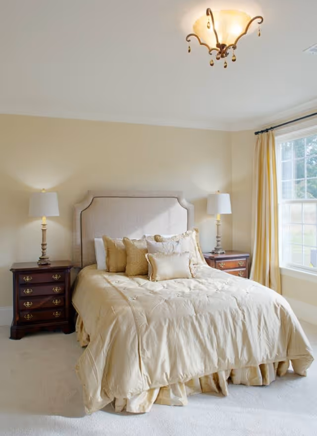 A neatly made bed with a beige comforter and multiple decorative pillows in a bedroom. The bed has a padded headboard and is flanked by two wooden nightstands, each with a table lamp. There is a large window with beige curtains on the right side, allowing natural light to brighten the room.