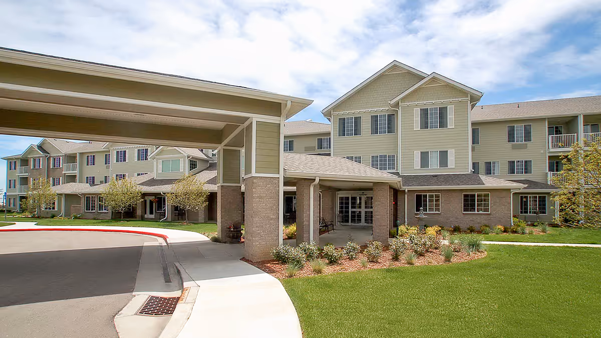 Front entrance of a multi-story senior living facility with a covered drop-off portico, landscaped lawn, and multiple apartment windows.