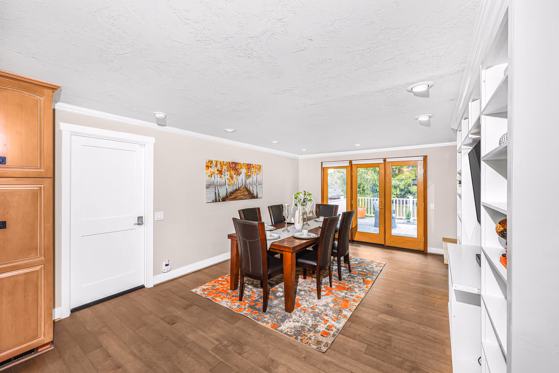 A bright dining room with a wooden dining table set for six people with dark brown chairs. The room has light-colored walls, a colorful area rug under the table, a painting of a tree-lined path on the wall, and large wooden-framed glass doors leading outside. There is a white built-in shelving unit on the right and wooden cabinetry on the left.