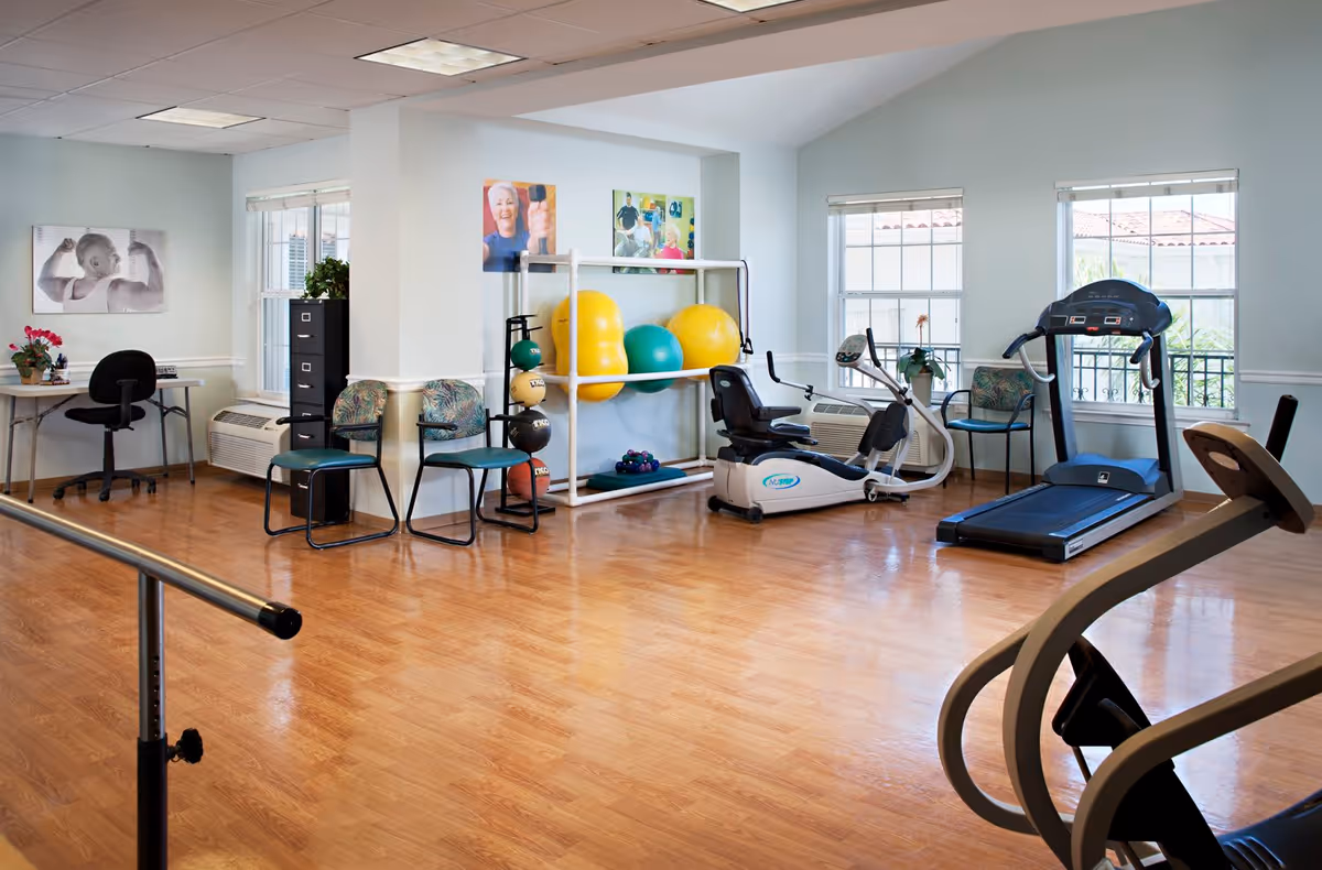 A bright exercise room with wooden flooring featuring a treadmill, a recumbent exercise bike, a rack with large colorful exercise balls, and a set of dumbbells. There are two chairs and a small desk with a chair in the corner. The room has large windows letting in natural light and motivational photos on the walls.