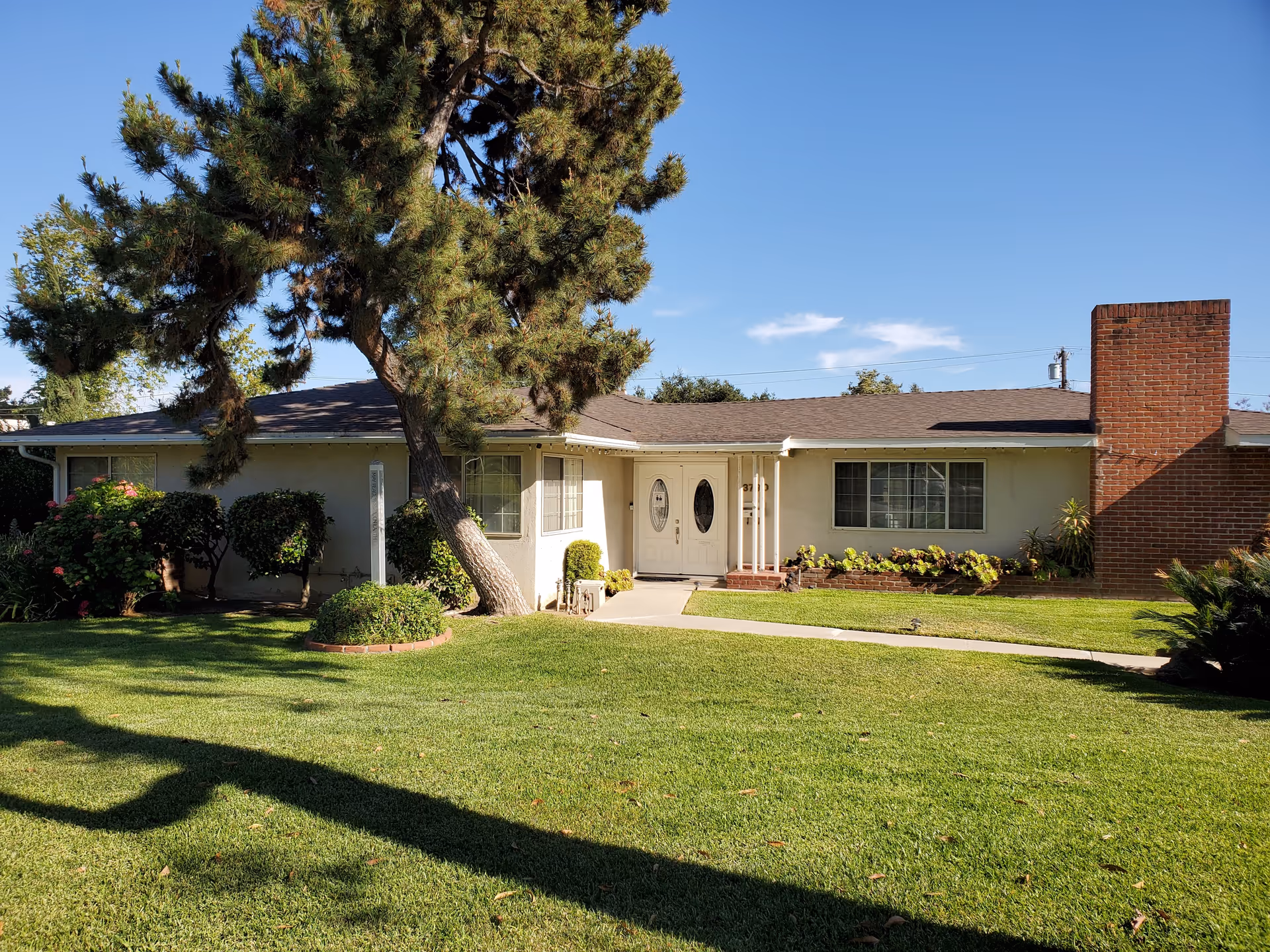 Single-story house front with a manicured lawn, a leaning tree, double front doors and a brick chimney.