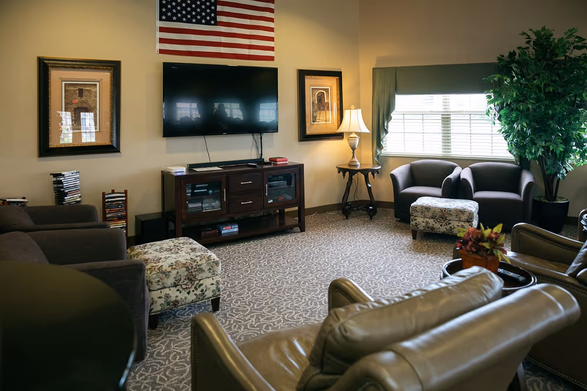 A cozy senior living common area with several armchairs and floral ottomans arranged around a television mounted on the wall. The room features a patterned carpet, framed artwork, a table lamp on a small side table, a large window with blinds and green valance, a potted plant, and an American flag hanging above the TV.