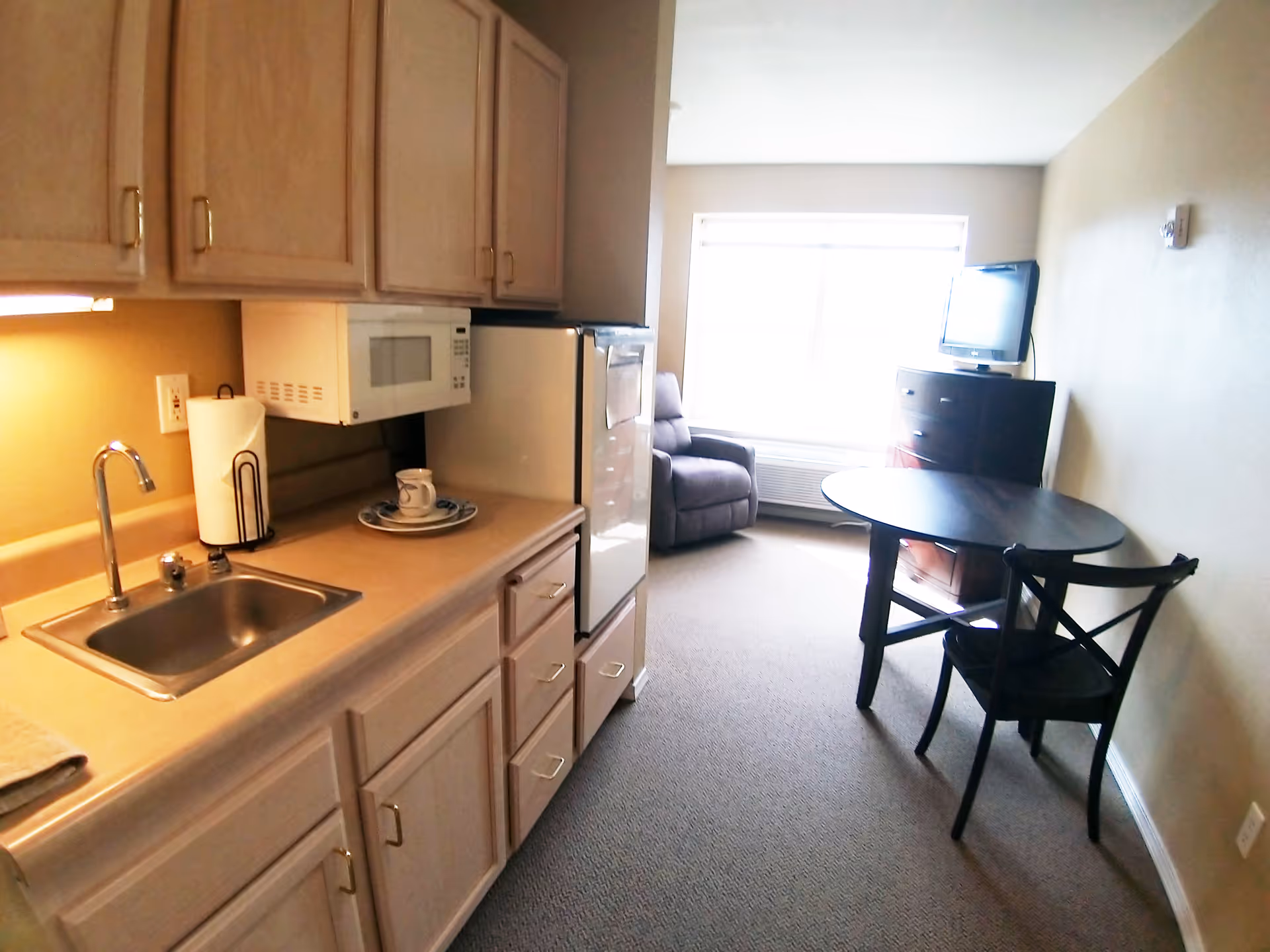A small senior living apartment kitchenette with light wood cabinets, a sink, microwave, and mini refrigerator on the left. On the right side, there is a round dark wood table with two chairs, a recliner chair near a large window, and a dresser with a flat-screen TV on top.