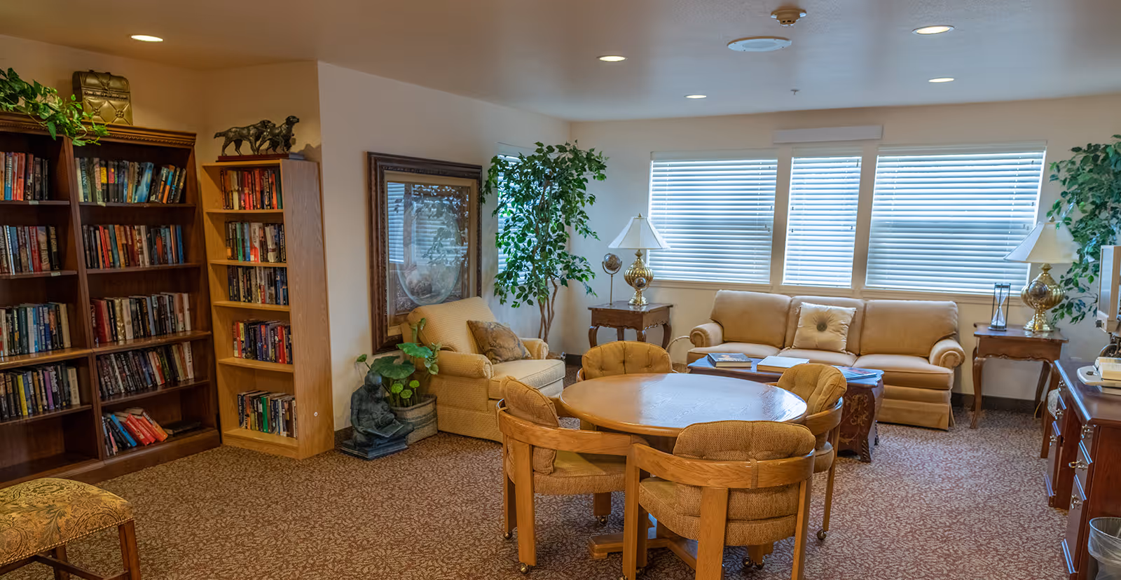 A cozy living room area in a senior living facility featuring a round wooden table with four cushioned chairs on wheels, a beige sofa, an armchair, two side tables with lamps, large windows with blinds, two bookshelves filled with books, a framed artwork on the wall, and several potted plants.