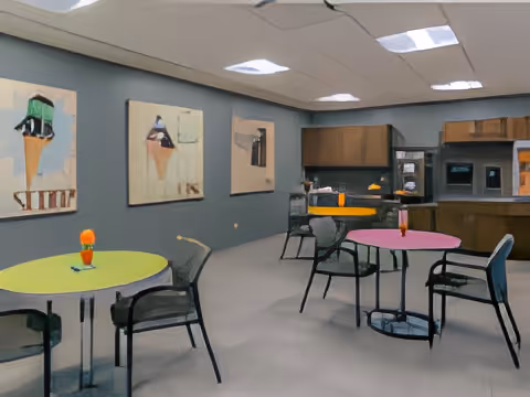 Interior view of a communal dining area with round tables covered in colorful tablecloths, each with a small flower vase. Several chairs are arranged around the tables. The room has a kitchen area in the background with cabinets, a microwave, and a refrigerator. Abstract paintings hang on the wall.