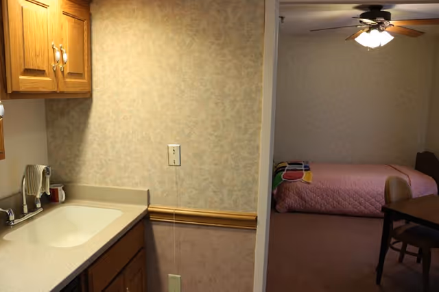 Interior view of a small kitchenette with a sink and cabinets leading into a bedroom with a pink bedspread and ceiling fan.