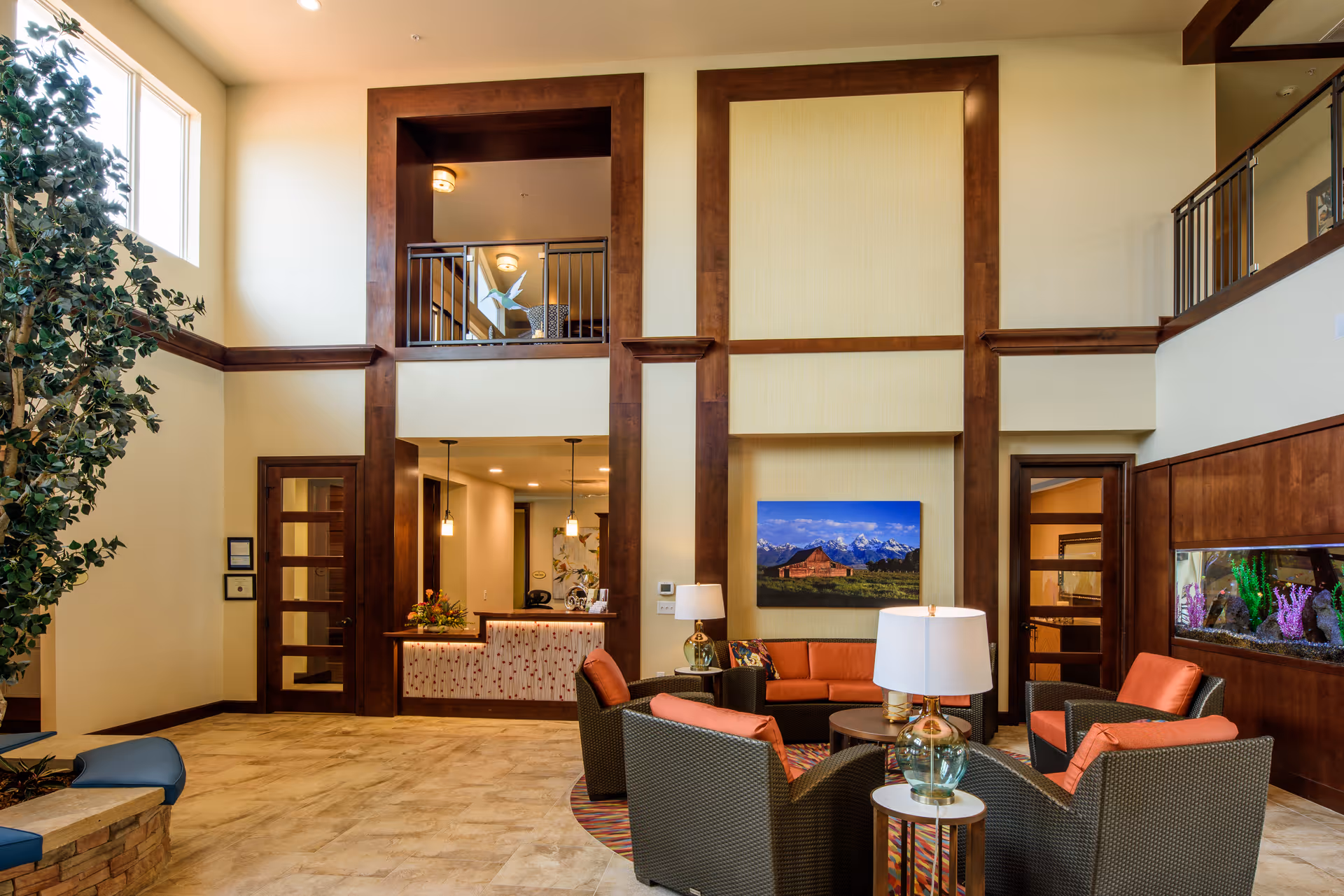 A spacious and well-lit assisted living facility common area with high ceilings, large windows, and wood trim accents. The seating area includes wicker chairs with orange cushions arranged around a small round table with a lamp. There is a large aquarium built into the wall on the right and a colorful landscape painting on the back wall. A reception desk is visible in the background with pendant lights hanging above it. A large indoor plant is on the left side near the windows.