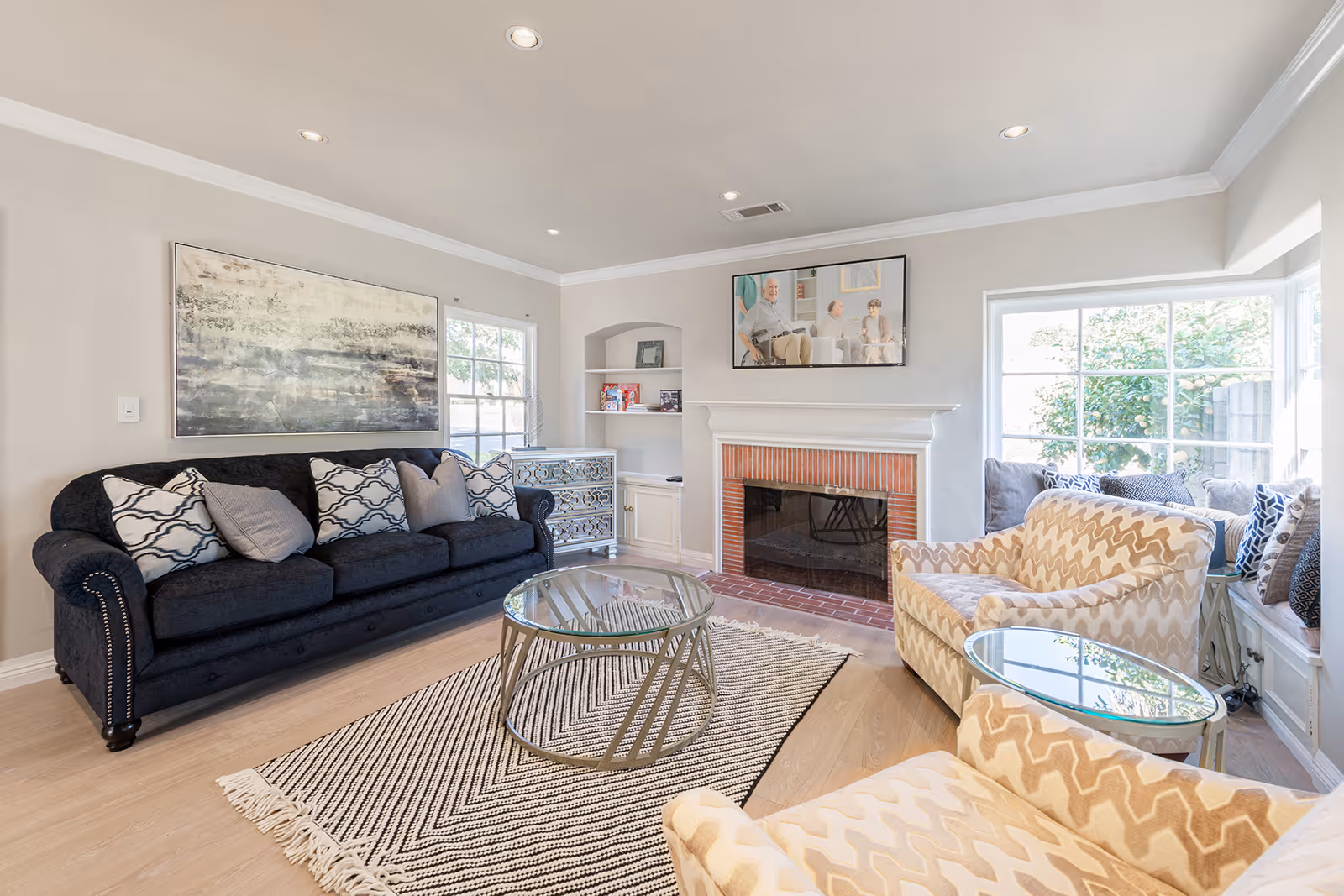 A bright and cozy living room with a black sofa adorned with patterned pillows, two beige armchairs with a zigzag pattern, a glass coffee table on a striped rug, a fireplace with a brick surround, built-in shelves, and large windows letting in natural light.