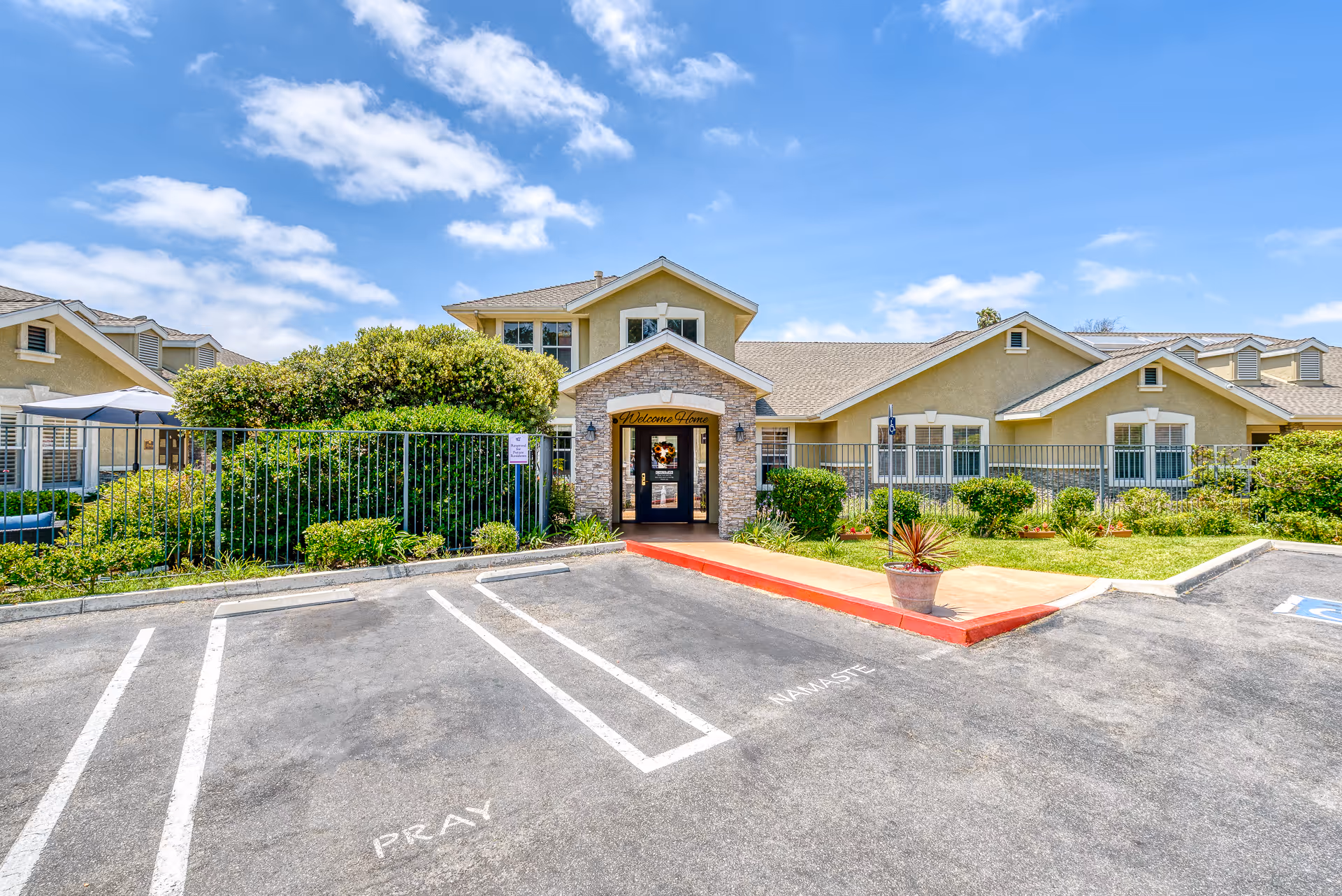 Entrance of a single-story senior living building with a stone archway, landscaped front lawn, and empty parking spaces under a blue sky.