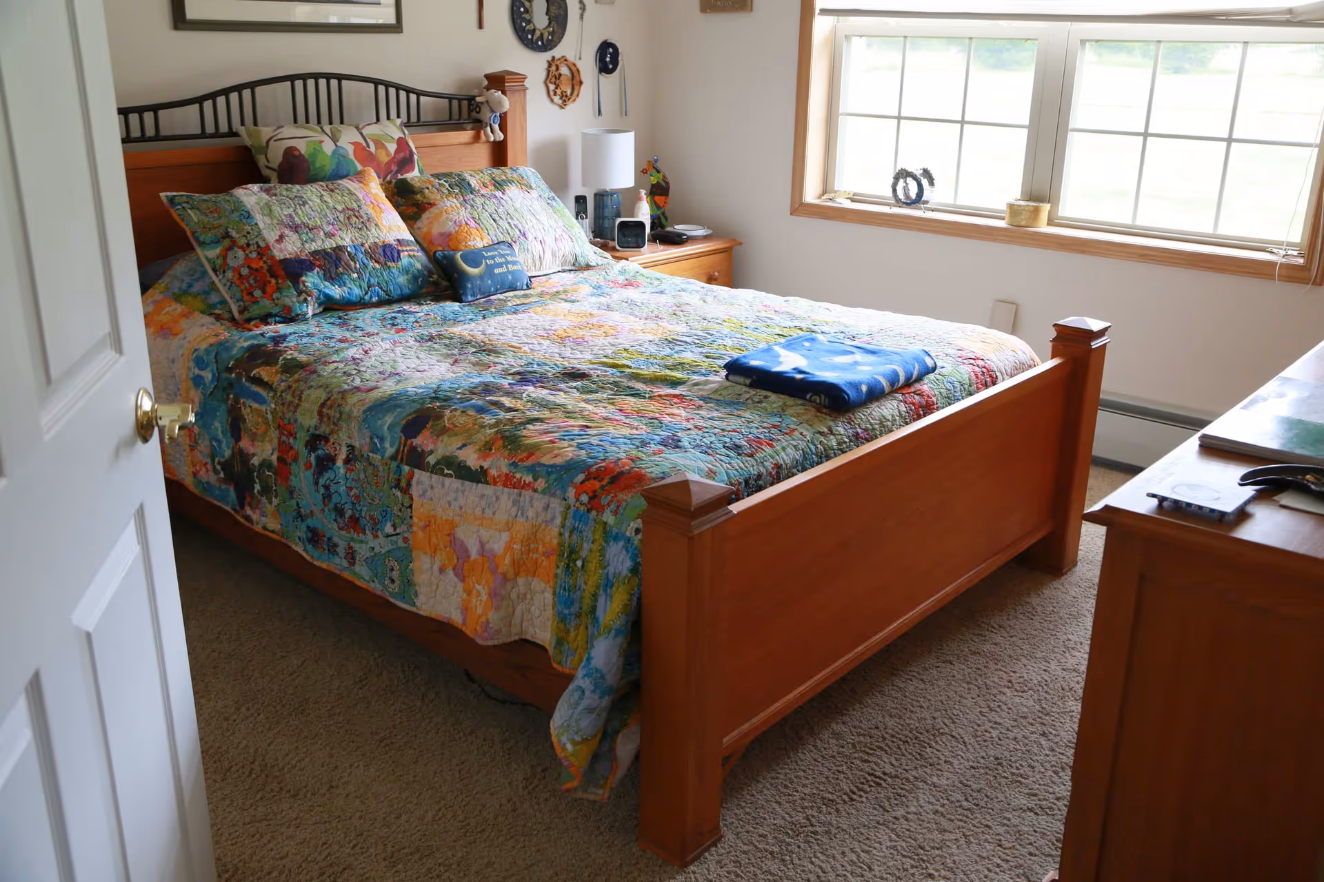 A cozy bedroom with a wooden bed frame and a colorful patchwork quilt on the bed. There are multiple pillows, including one with a bird design and a small decorative pillow with text. A wooden nightstand beside the bed holds a lamp, a clock, and some small items. A large window with a wooden frame lets in natural light, and a wooden dresser is partially visible on the right side of the image.