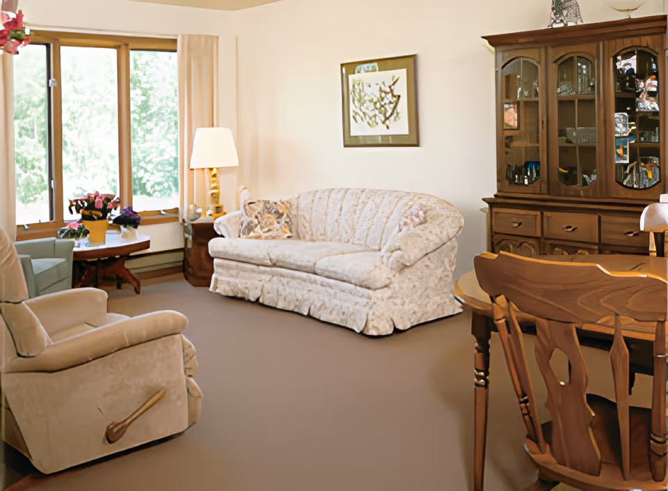 Bright living room with a floral sofa, recliner, wooden dining table and china cabinet beside large windows.