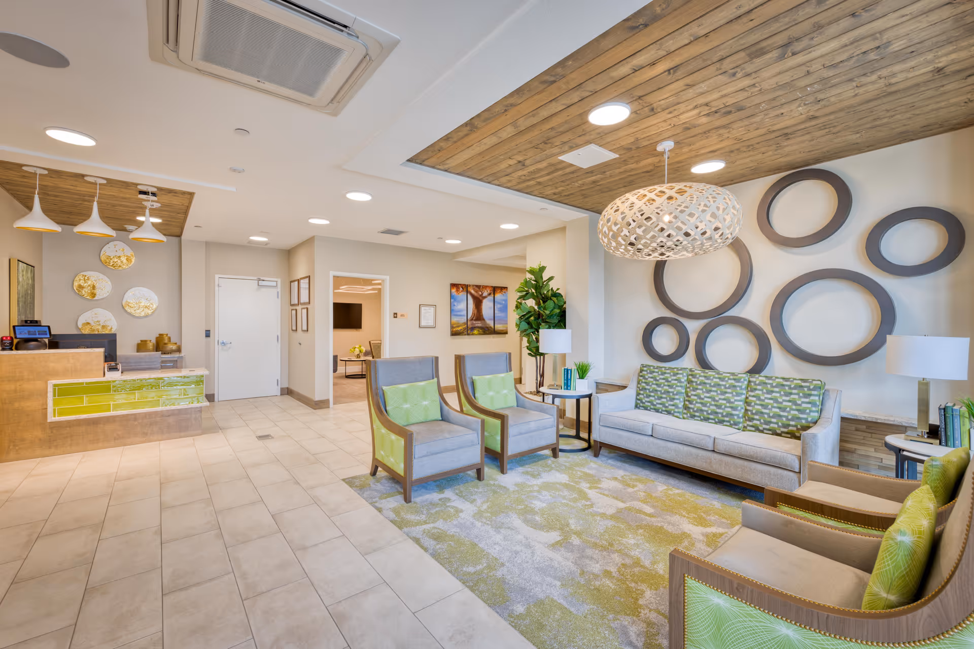 A bright and modern senior living facility lobby with a reception desk on the left featuring green tile accents. The seating area on the right includes a gray sofa with green patterned cushions, two matching armchairs, and two additional chairs with green cushions, all arranged on a green and gray patterned rug. The walls are decorated with circular wall art and a large woven pendant light hangs from a wooden ceiling panel. There is a plant and side tables with lamps and books, and a hallway with framed pictures is visible in the background.