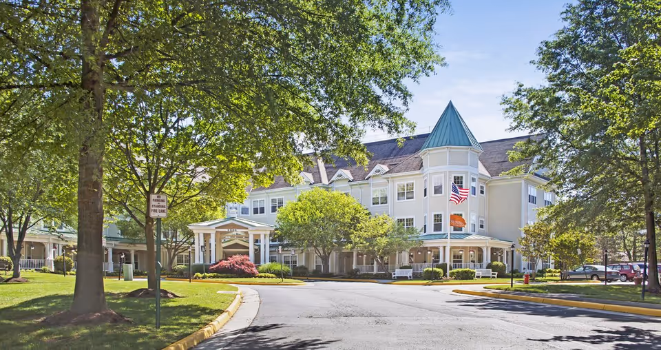 Front exterior of a large three-story senior living building with a turret, American flag, driveway, and landscaped trees and lawn.