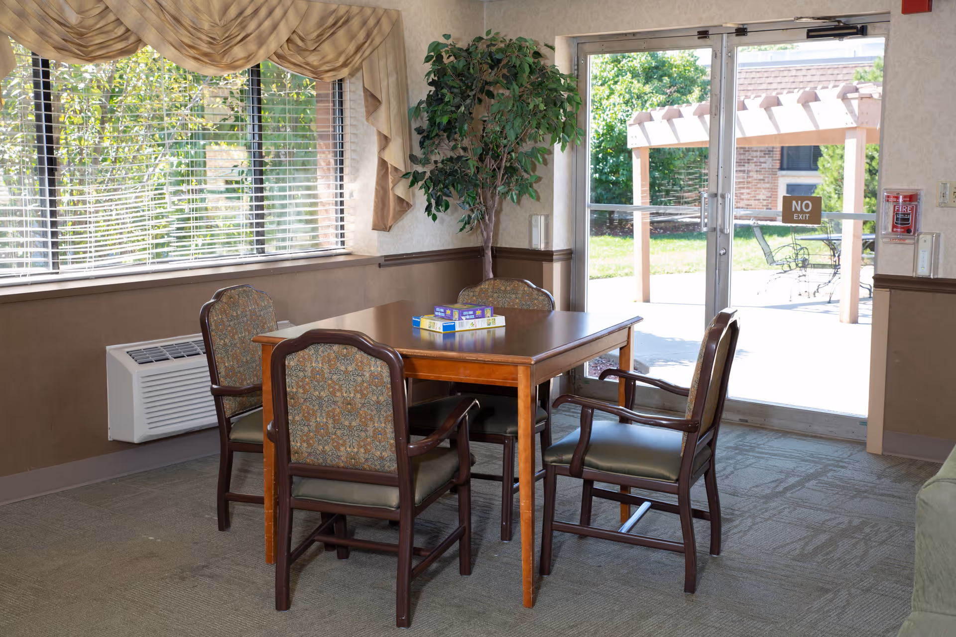 A small seating area inside a room with a wooden table and four upholstered chairs. On the table, there are two board games. The room has a large window with blinds and beige curtains, a potted plant in the corner, and a glass door leading outside to a patio with a pergola and outdoor furniture. A fire alarm is mounted on the wall near the door.