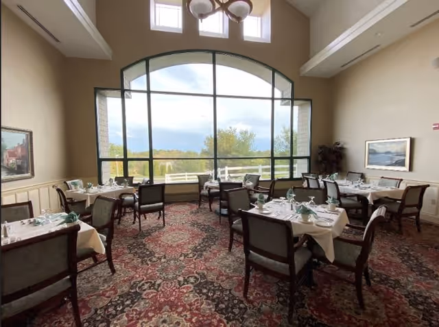 Dining room with multiple tables covered in white tablecloths, set with plates, glasses, and folded green napkins. Large arched window with a view of trees and sky outside. The room has high ceilings, a patterned carpet, and framed artwork on the walls.
